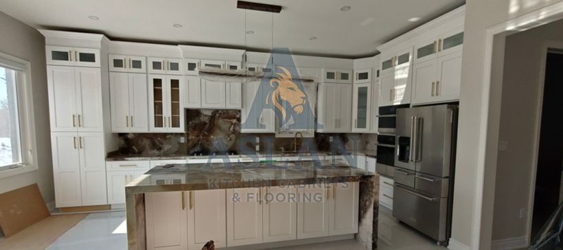 White shaker kitchen with glass-front uppers, gold pulls and a natural stone waterfall island in daylight