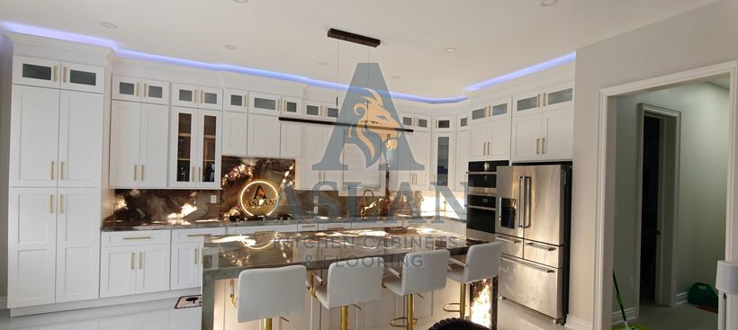 White shaker kitchen at night with cove lighting, a backlit natural stone backsplash and waterfall island with gold bar stools